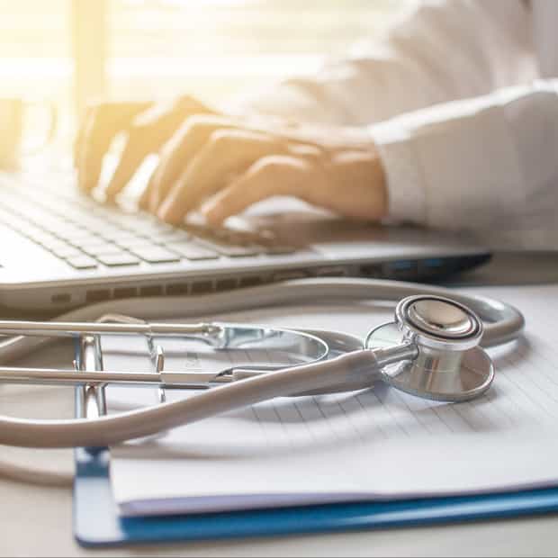 Primo piano di un medico che digita con concentrazione su un laptop, con uno stetoscopio in primo piano, a sottolineare l’atmosfera medica.