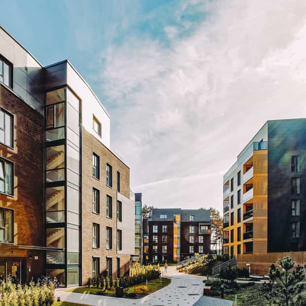 Complesso residenziale moderno con due edifici a più piani separati da un sentiero lastricato, circondato da cespugli e alberi, sotto un cielo sereno.