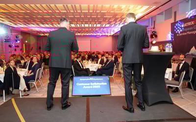 View from a stage during a formal conference event. Two people stand at the front facing a seated audience at round tables. A screen in front of the stage displays the text “European Partner Conference – Customer Satisfaction Award 2025.” Stage lighting and table settings are visible.