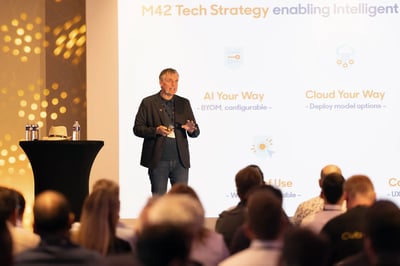Conference presentation scene with a person standing on a stage in front of a large slide. The slide shows the heading “M42 Tech Strategy enabling Intelligent” and sections such as “AI Your Way” and “Cloud Your Way.” An audience is seated in the foreground.