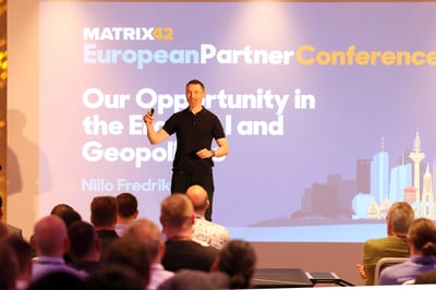 A conference scene with a person standing on a stage in front of a large screen. The screen displays the text “Matrix42 European Partner Conference” and a presentation title. An audience is seated in the foreground.
