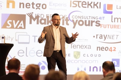 A conference scene with a person standing on a stage and gesturing with both hands. Behind, a large screen displays various company logos and partner names. Audience members are visible seated in the foreground.