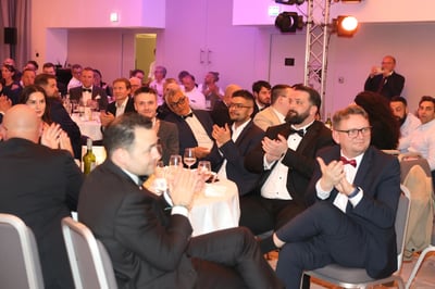 People seated at round tables during a formal event indoors. Attendees wear formal attire, with glasses, bottles, and table settings visible. Stage lighting and additional guests appear in the background.