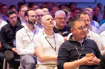 Audience seated in rows at a conference, listening to a presentation. Several attendees wear lanyards, glasses, and casual business clothing. The background shows additional participants under stage lighting.