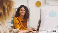 A smiling woman wearing glasses and an orange blouse is sitting at a desk in an office environment, looking at a computer screen. In the foreground, another person is sitting, slightly blurred.