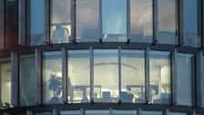 View of a modern office building with large curved glass windows. Inside, several people are working at desks in a brightly lit environment in the evening or at night.