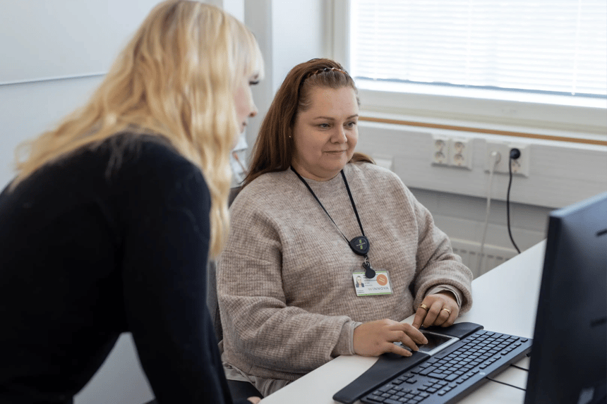 Two individuals in a professional setting, one seated at a computer wearing a badge labeled 'WINNOVA,' and the other standing nearby, engaged in a collaborative task.