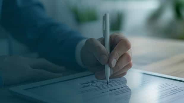 A hand holding a stylus pen signing a digital document on a tablet, representing modern contract management in a professional setting.