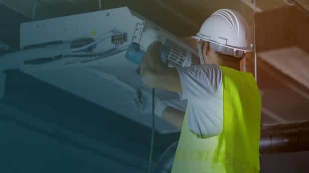 A worker in a neon yellow safety vest and white hard hat performing maintenance on a ceiling-mounted HVAC unit in an industrial setting, representing facility management.