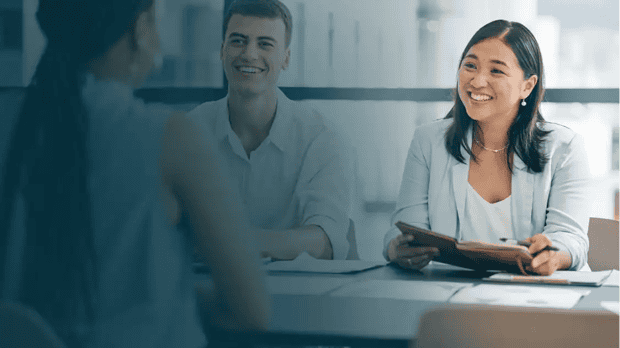 Three people in a professional Human Resources meeting, with a woman in a light blue blazer smiling and holding a notebook, a man in a light shirt smiling, and another woman with braided hair facing away, in a bright office setting.