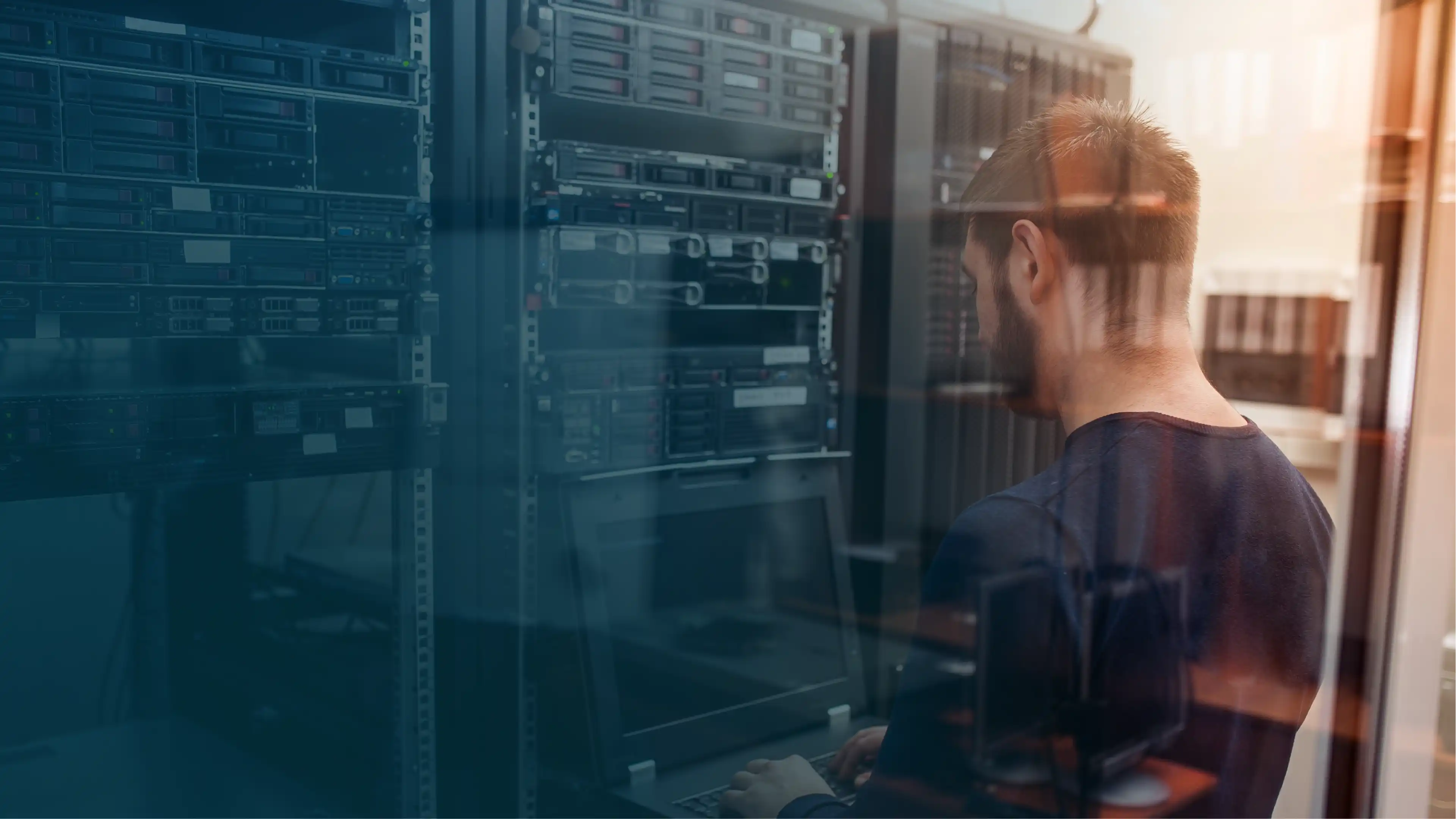 A person working on a laptop in a data center, facing server racks filled with networking equipment, viewed through a glass barrier with warm lighting and reflections.