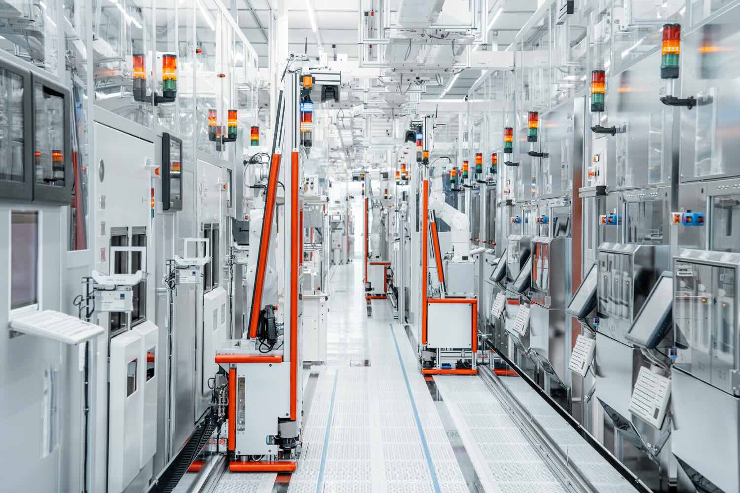 Interior view of an automated manufacturing facility with machines and robotic units aligned along a central corridor. Control panels, signal lights, and cabling are visible throughout the clean, well‑lit production area.