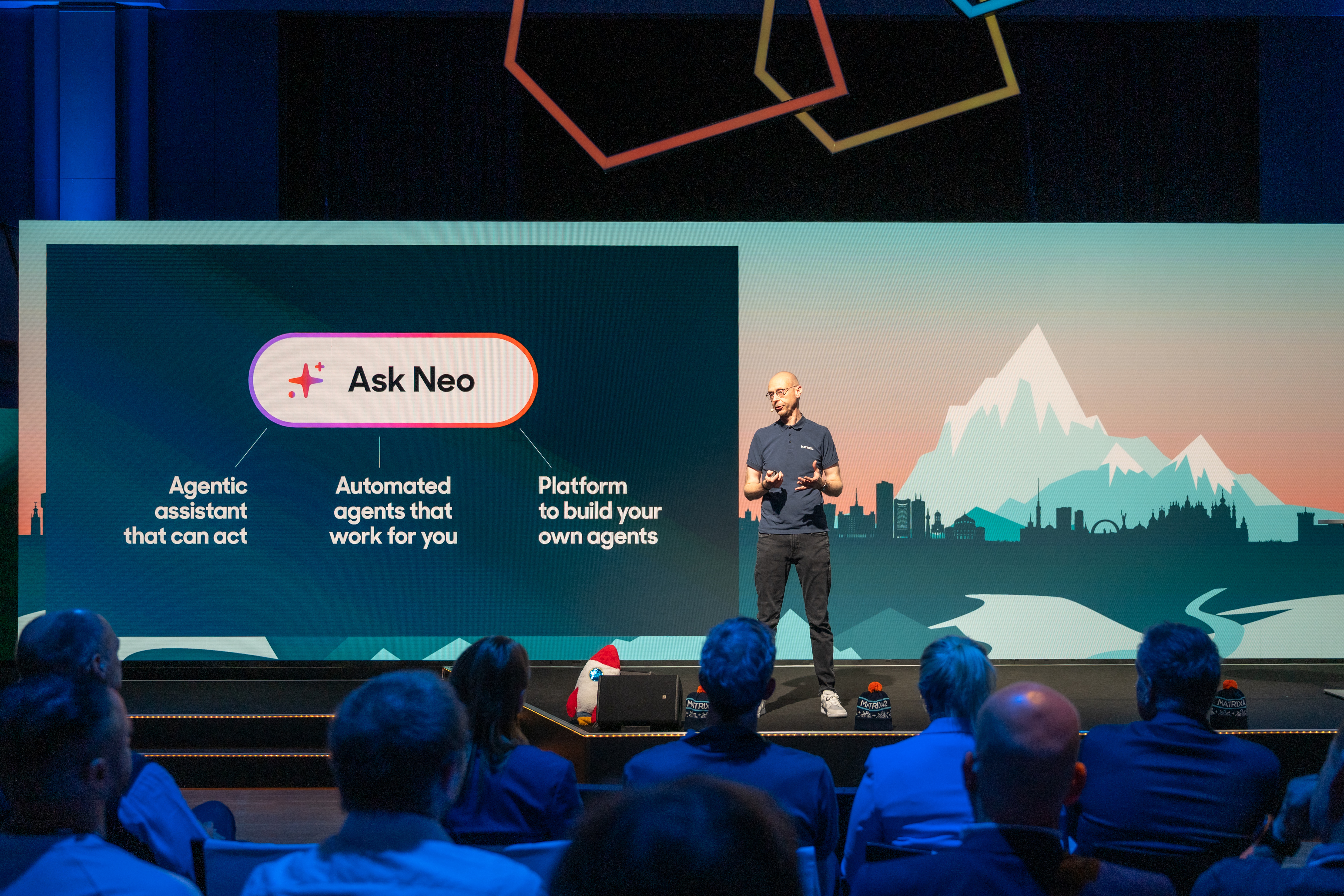 Speaker presenting Matrix42 Neo on stage, with a slide showing Ask Neo: an agentic assistant that can act, automated agents that work for you, and a platform to build your own agents