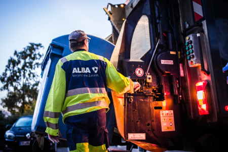 A sanitation worker in high‑visibility clothing operates the rear loading mechanism of a garbage truck. The vehicle shows control panels, warning lights, and safety labels, with another vehicle nearby in low light.