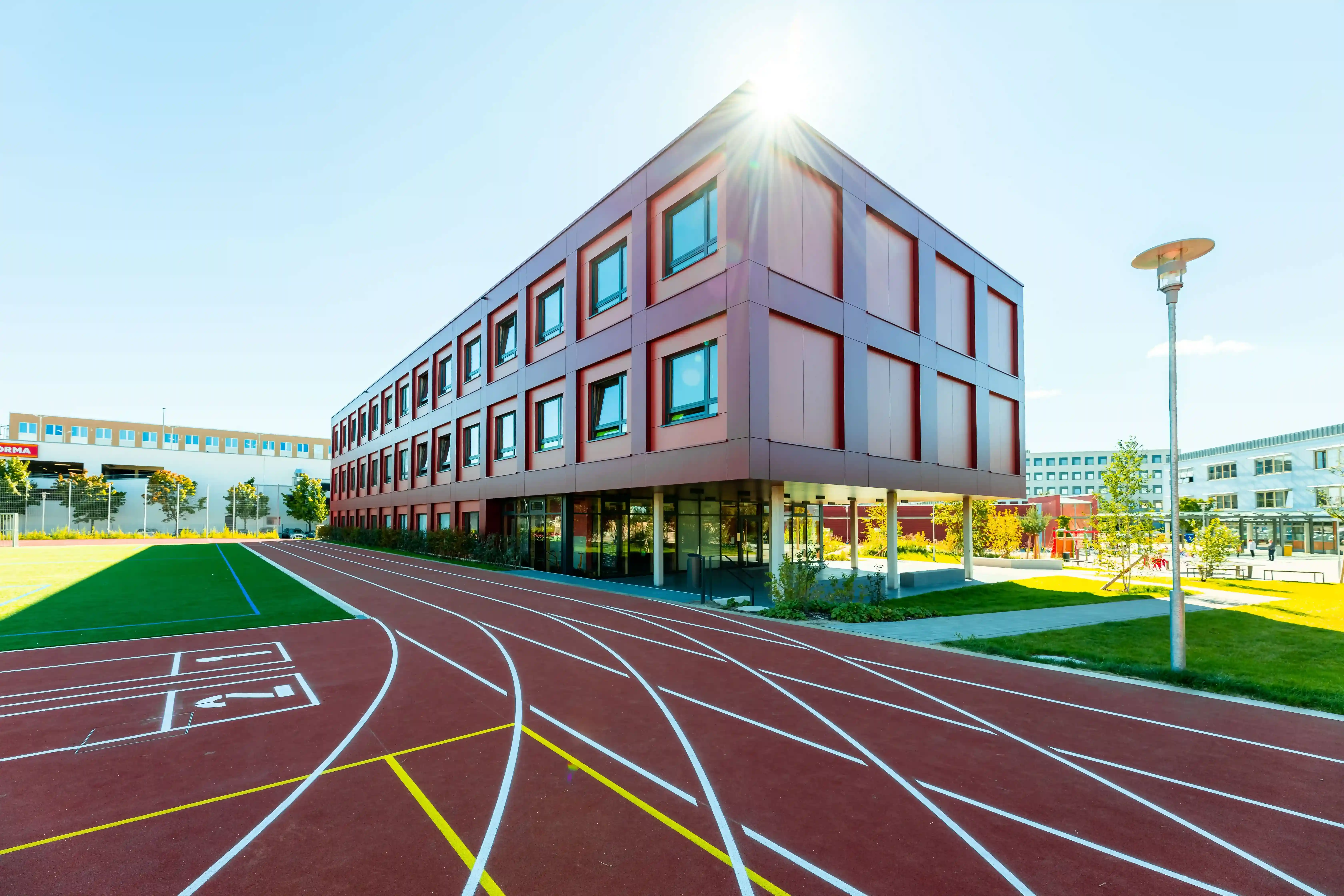 A modern three‑story building with rectangular windows stands beside a red running track with marked lanes. A pedestrian bridge and additional buildings are visible in the background under clear daylight.
