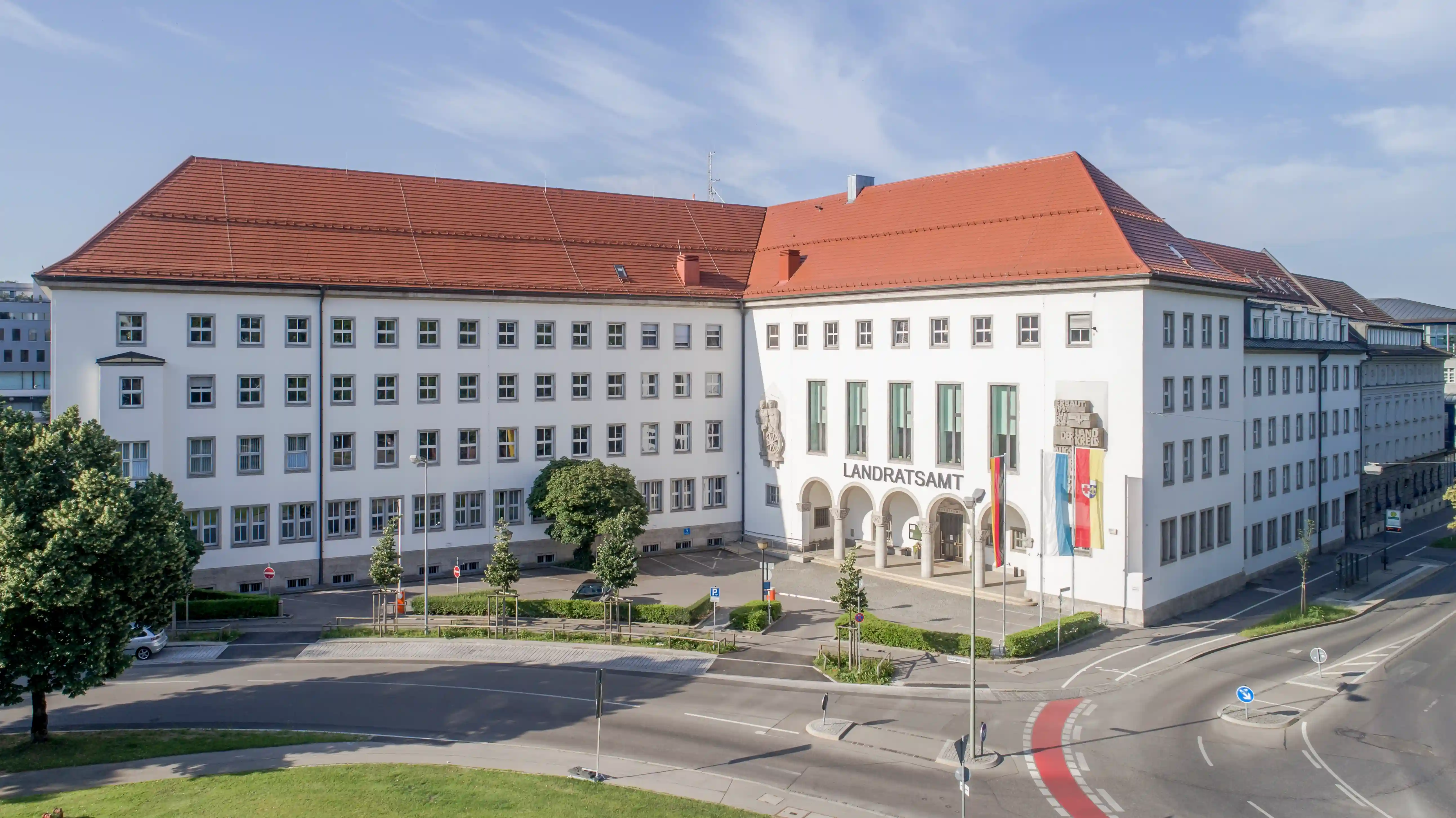 Außenansicht des Landratsamts Augsburg, eines großen weißen Gebäudes mit roten Ziegeldächern und mehreren Fensterreihen. Der Haupteingang liegt an einer Straßenkreuzung, davor stehen Bäume und Fahnen.