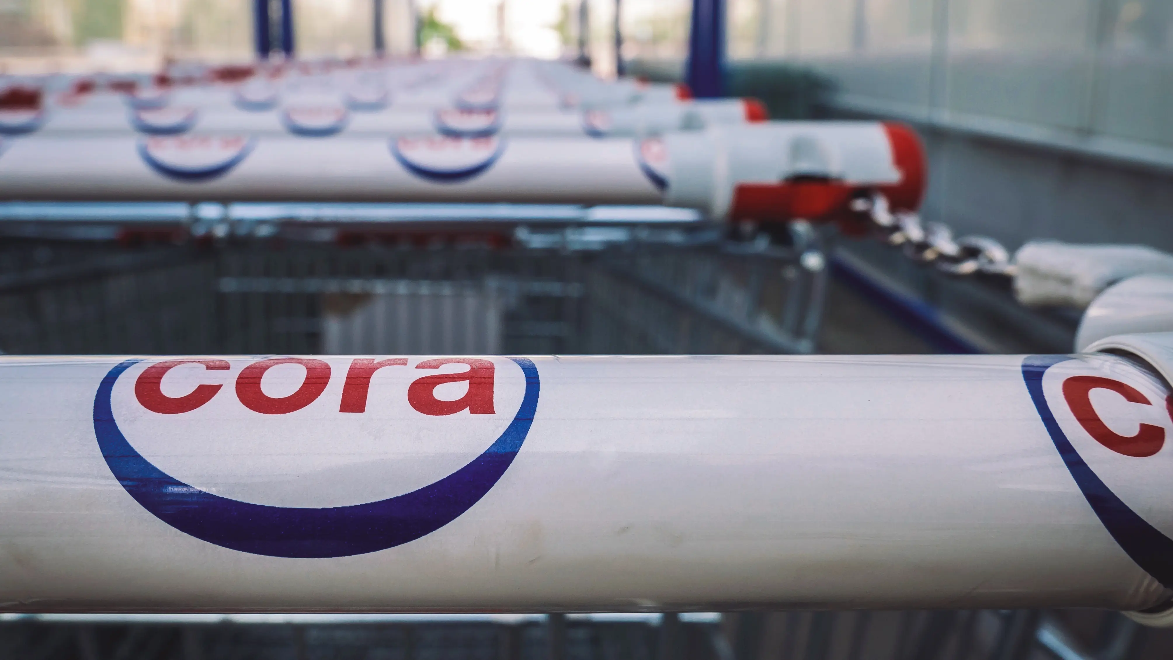 Close-up of shopping cart handles displaying the Cora logo in red and blue. Multiple rows of nested shopping carts extend into the background inside a retail store, with metal frames and soft indoor lighting visible.
