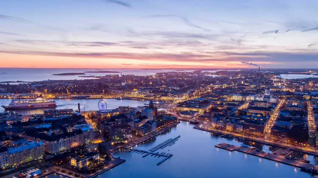 Ilmakuva Helsingistä auringonlaskun aikaan. Kuvassa näkyy valaistuja katuja, ranta-alueita, rakennuksia ja saaria. Etualalla näkyy satama veneineen ja maailmanpyörä, ja kaupunki jatkuu horisonttiin asti.