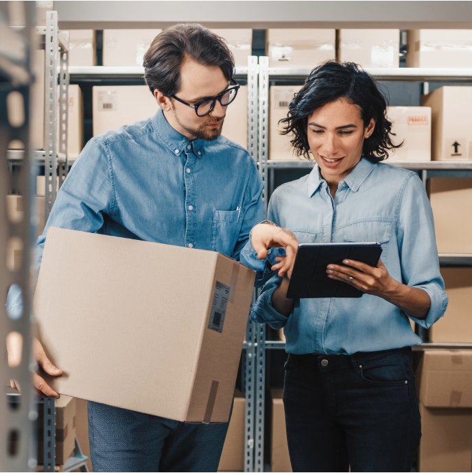 Two people stand in a storage or warehouse area with shelves of boxes in the background. One holds a large cardboard box while the other uses a tablet device. Both appear focused on information shown on the screen.