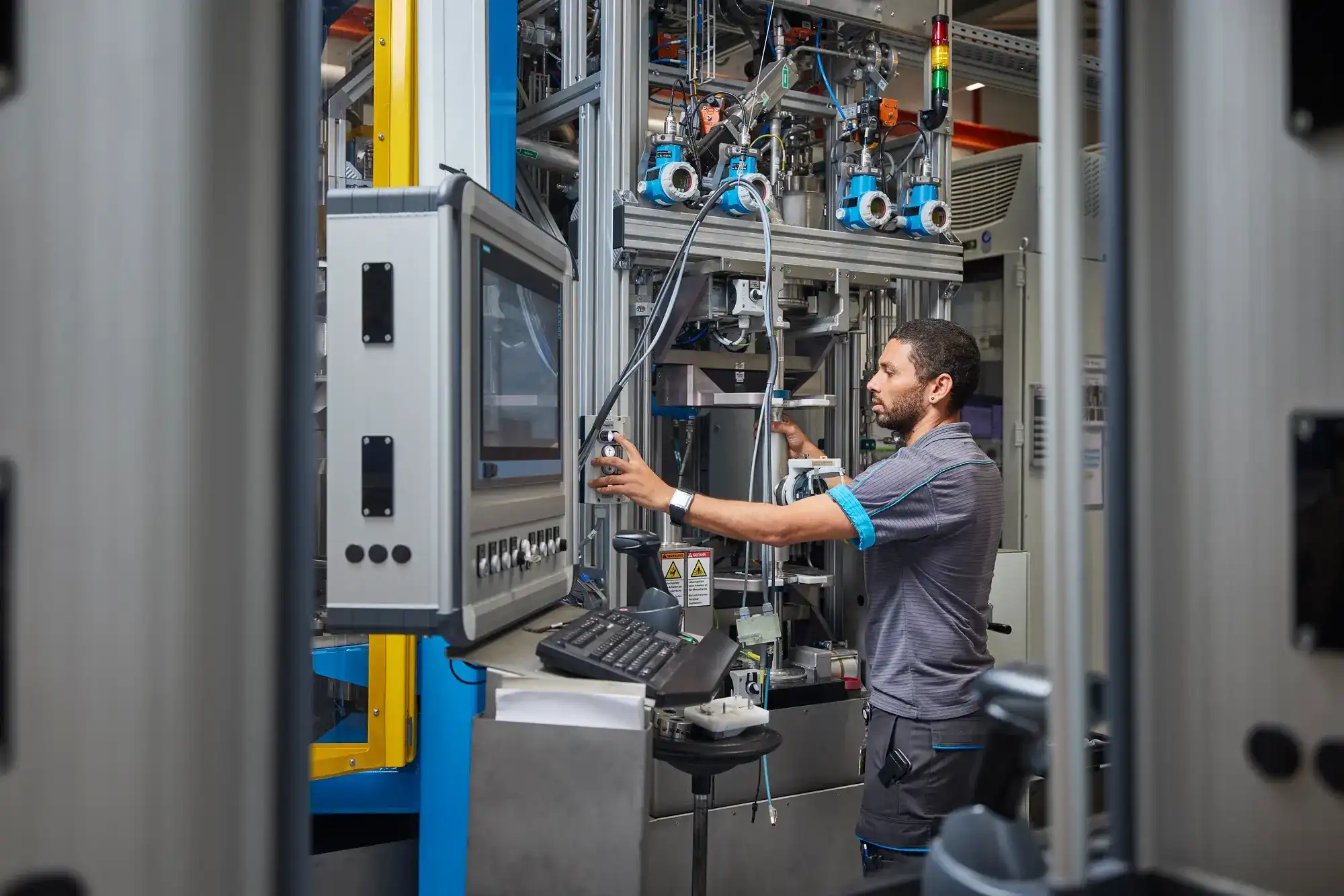 A person stands in an industrial setting operating machinery. A control panel with screen and keyboard is mounted on the left, while metal components, pipes, and robotic elements are visible around the workstation.