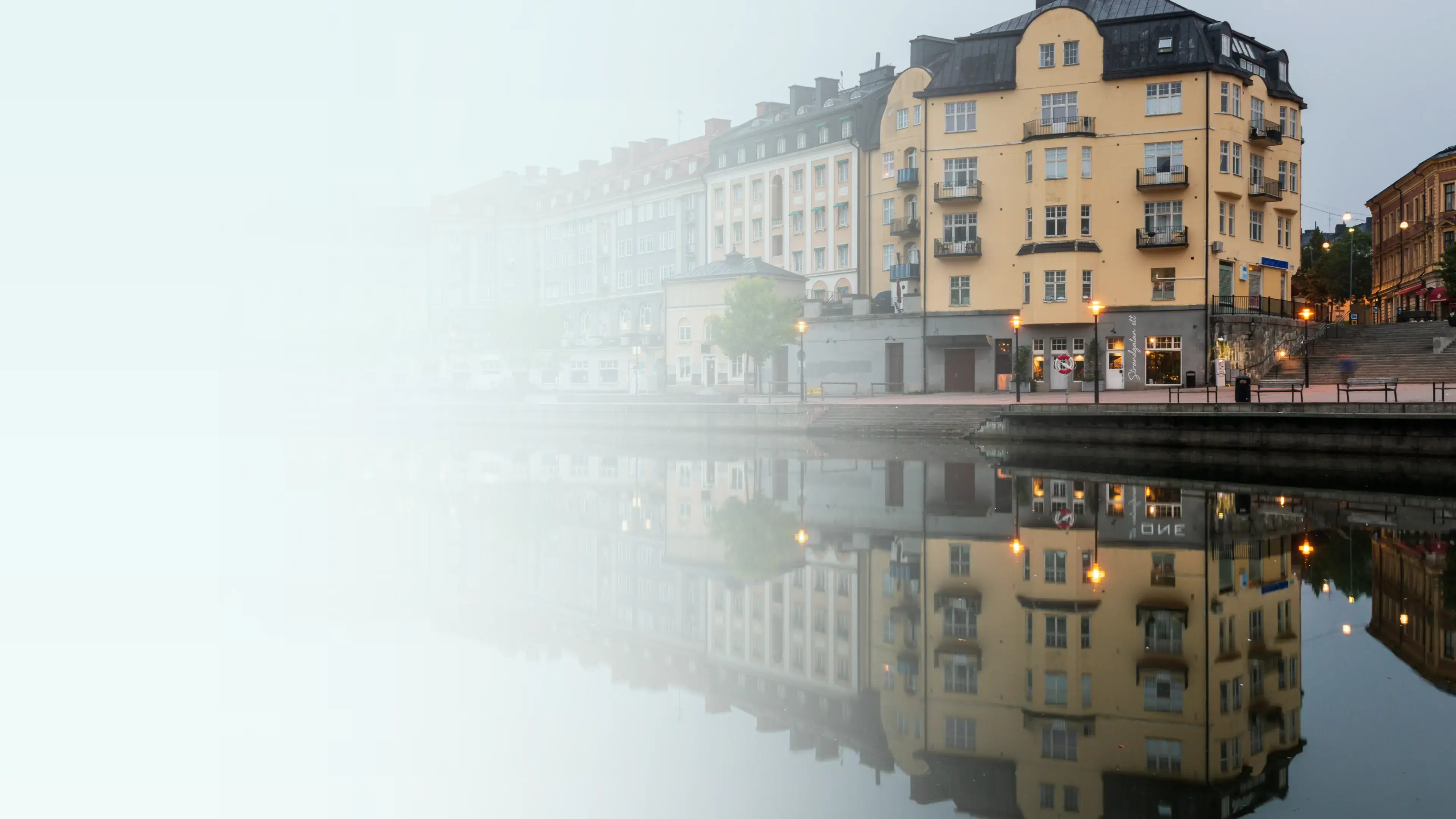 Vattennära vy i Södertälje med flerbostadshus som speglas i stilla vatten. Ljus längs kajen syns till höger, och bildens vänstra sida tonas ut i en ljus yta.