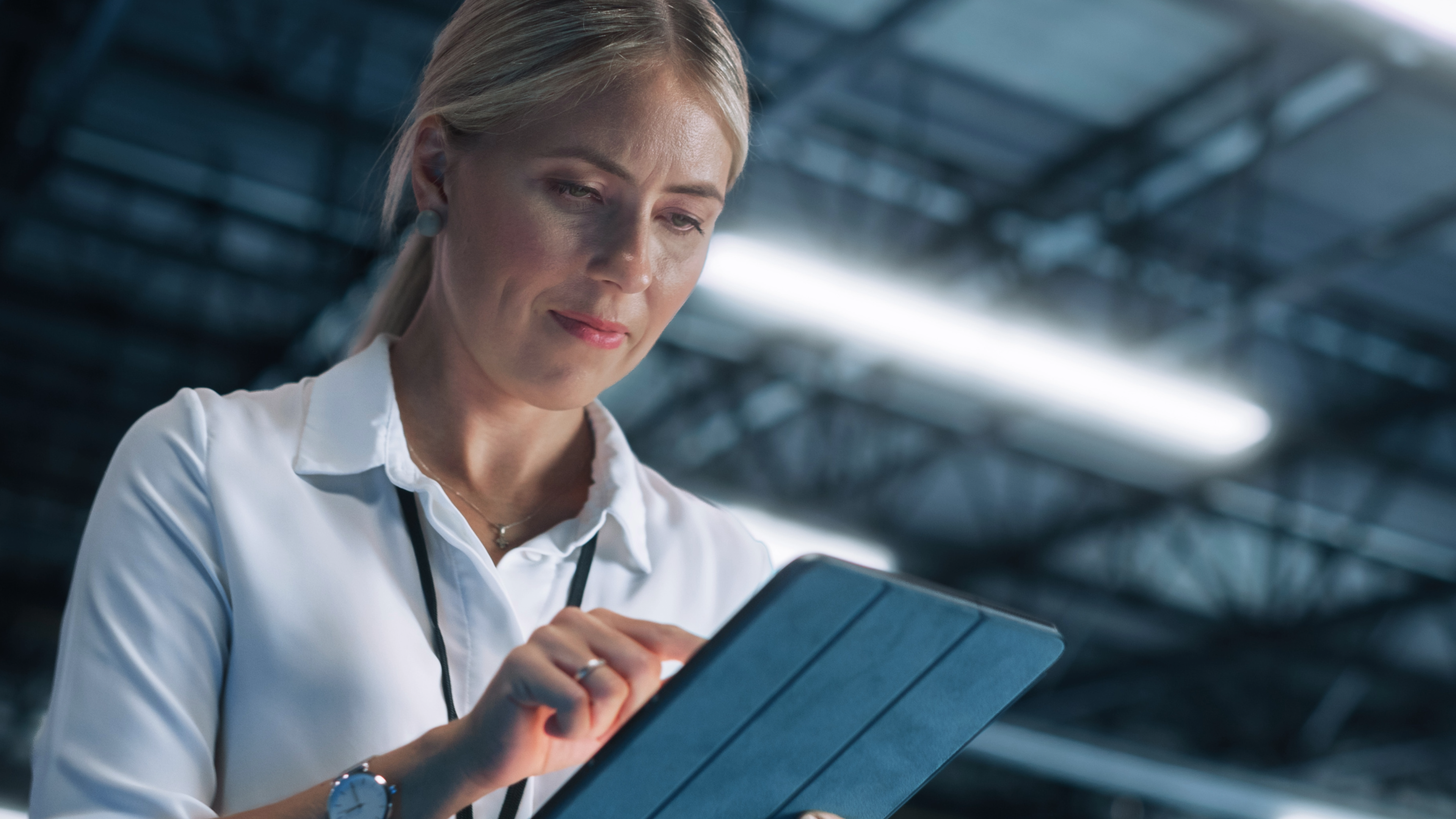 Eine Person steht in einem Innenraum und nutzt ein Tablet. Im Hintergrund sind Deckenbeleuchtung und Metallkonstruktionen zu sehen, die auf eine Industrie‑ oder Lagerumgebung hinweisen.