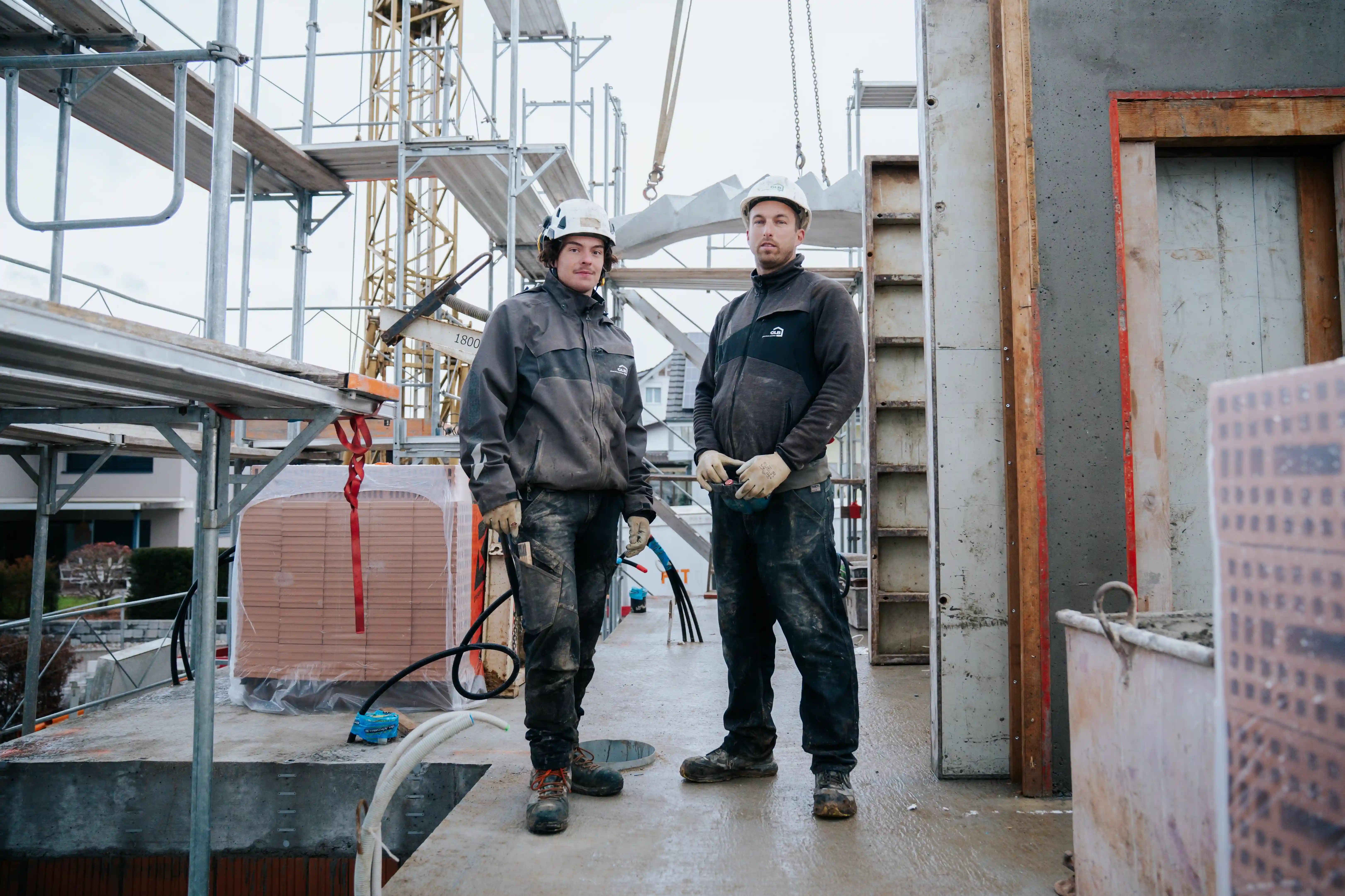 Two workers stand on a construction site platform surrounded by scaffolding, machinery, and building materials. Both wear safety helmets, gloves, and protective clothing. Metal structures, cables, and stacked bricks are visible, with partially built structures in the background.