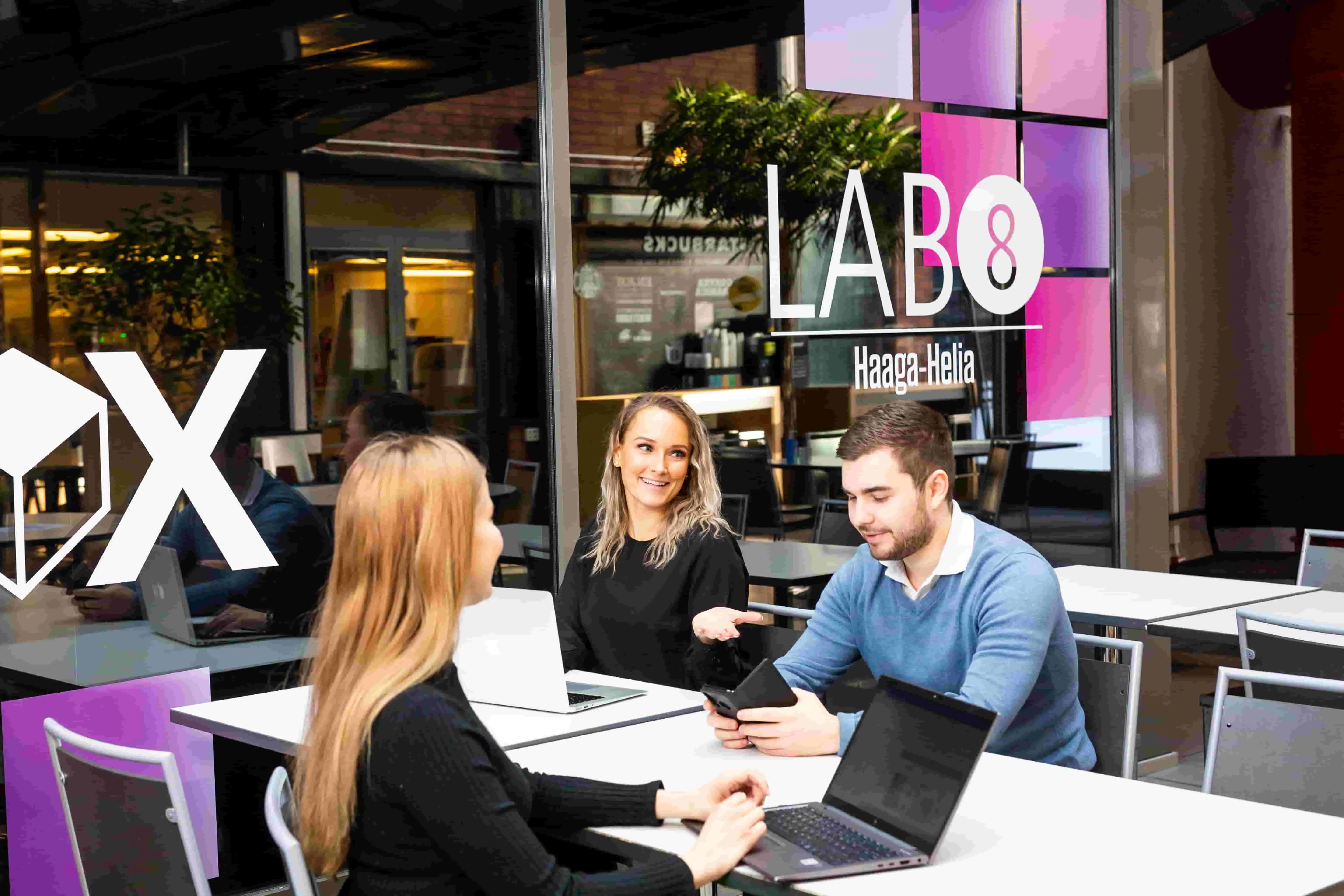 Three people sit around a table in a collaborative workspace with laptops and a mobile device. The glass wall behind them displays the text “LAB8 Haaga‑Helia.” Tables, chairs, and indoor plants are visible in the background.