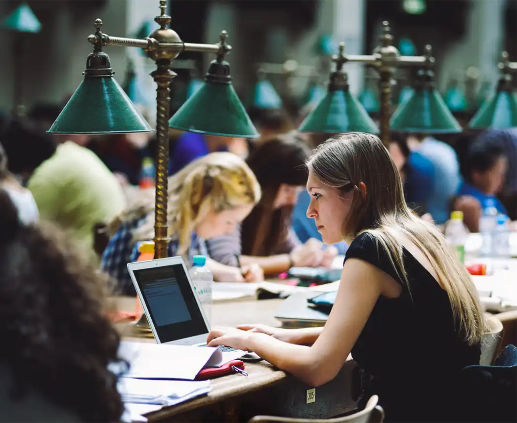 A student at the University of Vienna sits at a laptop in the library, with other students studying in the background.