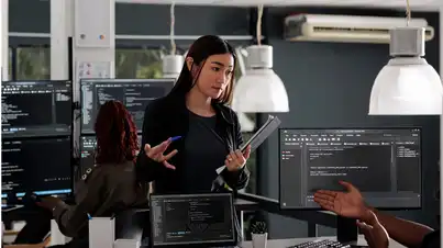 Several people work at computer stations with multiple monitors displaying code. One person stands between the desks holding documents and a pen, while another person gestures toward a screen. Large hanging lamps illuminate the workspace.
