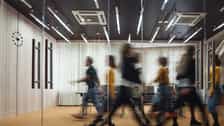 Blurred view of several people walking through a modern office corridor. Glass walls, doors, overhead lighting, and a wooden floor are visible, with motion blur emphasizing movement through the space.