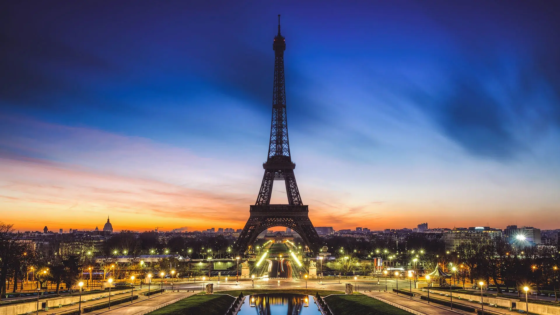 A view of the Eiffel Tower at dusk, seen from a distance with illuminated paths and reflections in a water basin. The sky shows a gradient from orange near the horizon to deep blue above.
