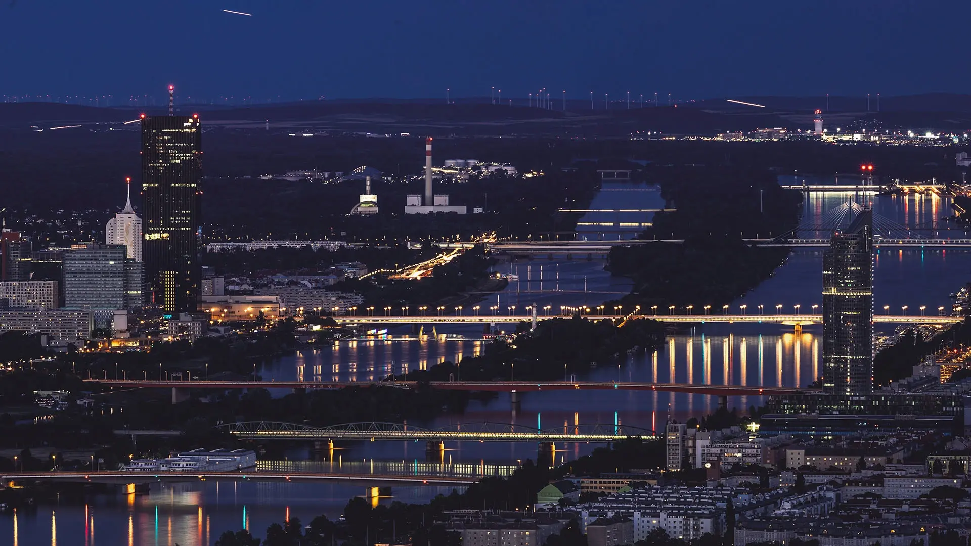 Nighttime view of Vienna with a river running through the city. Multiple illuminated bridges span the water, and buildings and towers are visible along the riverbanks under a dark sky.