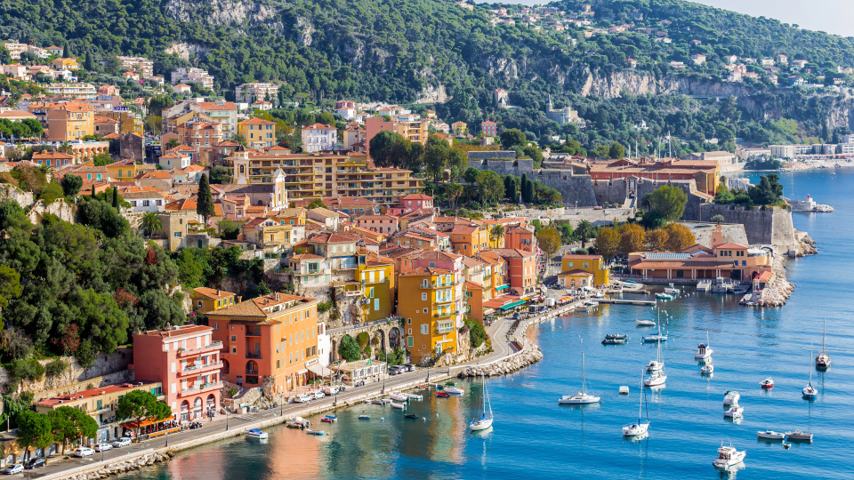 A coastal view of Nice, France, showing colorful buildings along the shoreline, hills covered with trees, and boats anchored in the harbor.