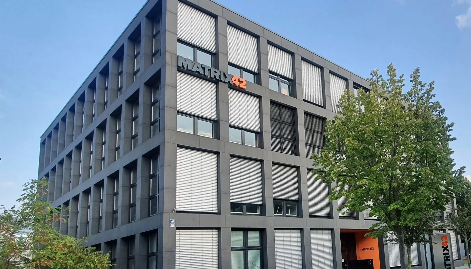 Exterior view of a modern office building with dark façades, large windows, and the Matrix42 logo mounted near the top. Trees stand in front of the building under a clear blue sky.