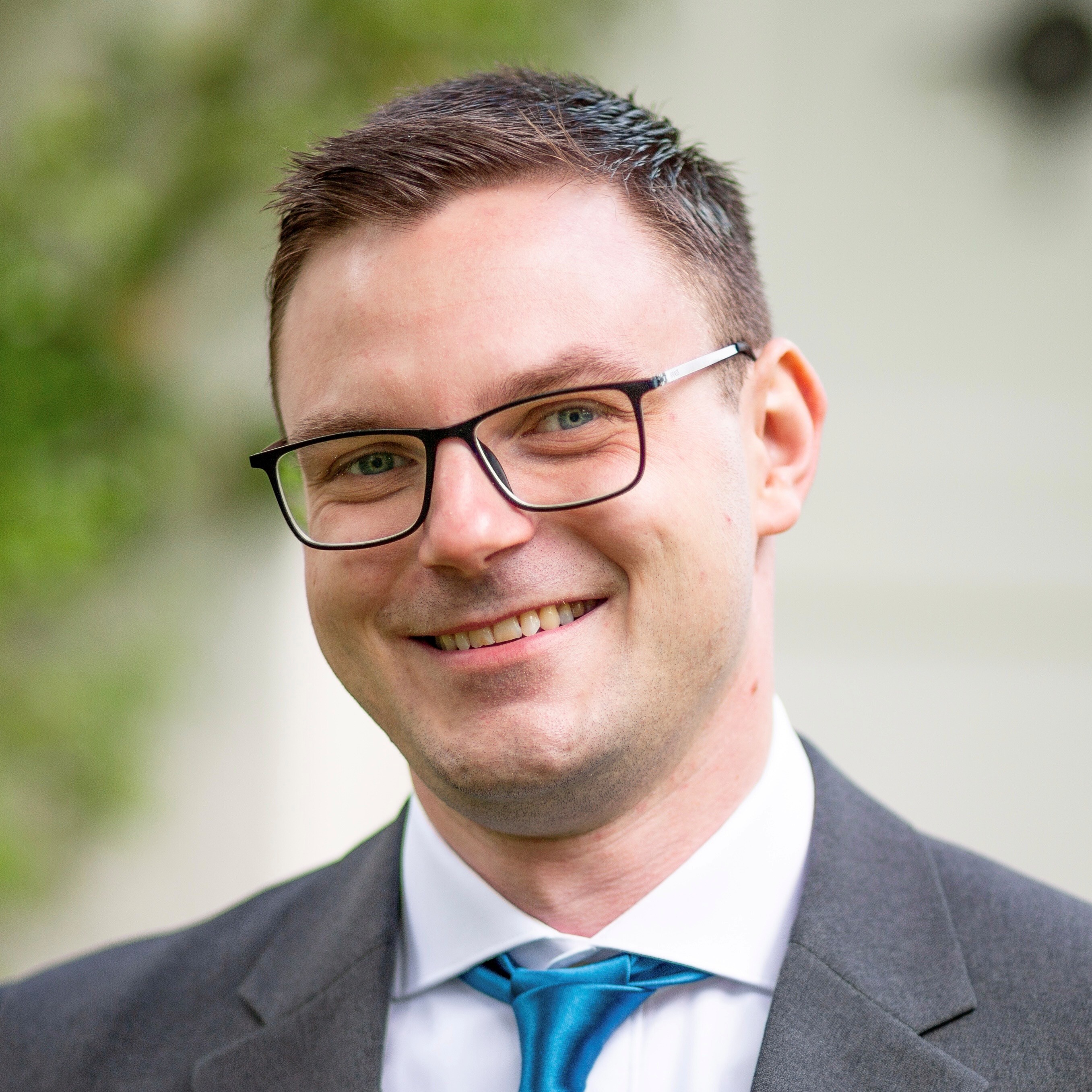 Portraitfoto einer Person mit einer Brille, die einen Anzug trägt und vor einem verschwommenen Hintergrund steht.