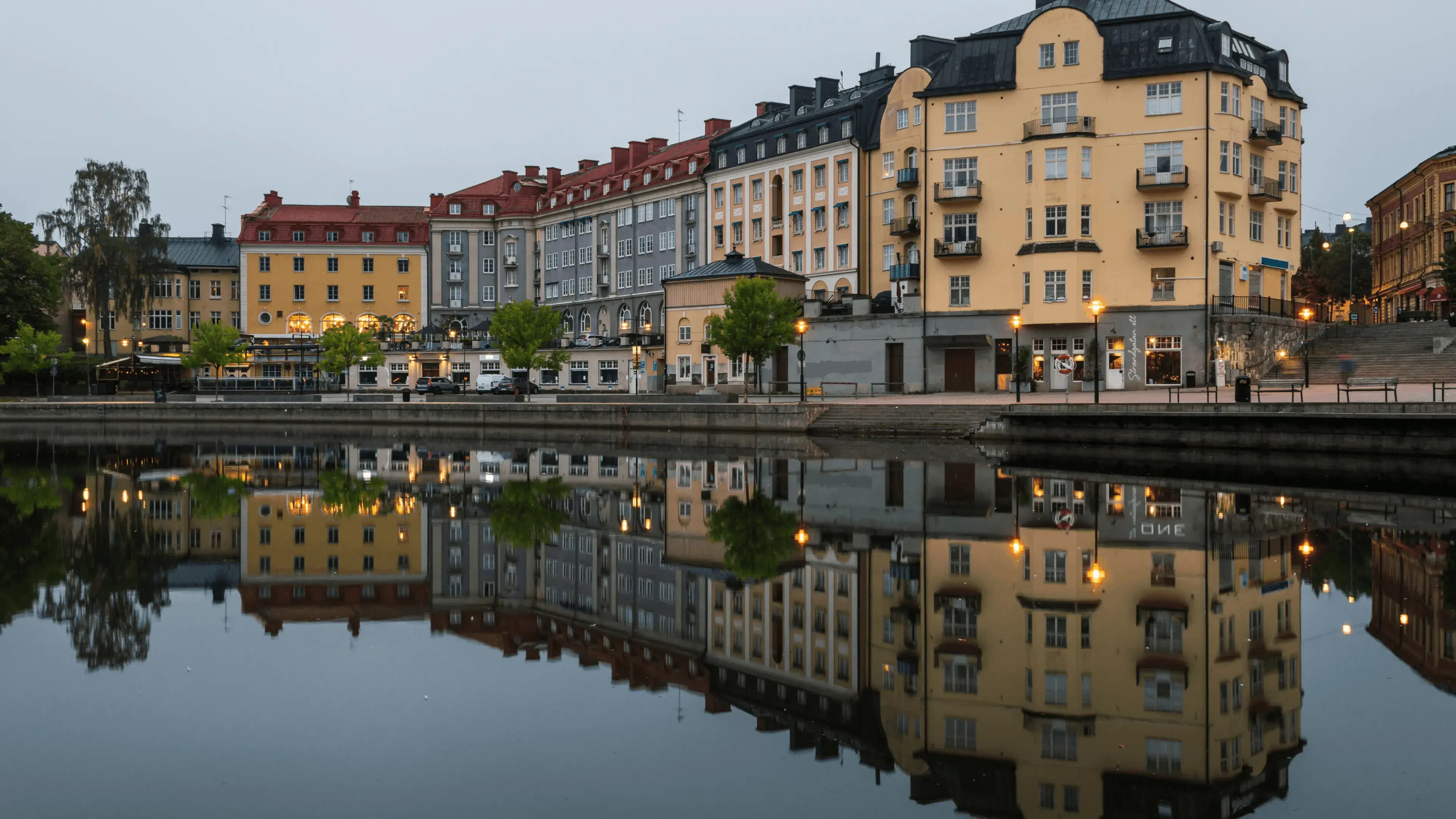 Vattennära vy med flerbostadshus och byggnader som speglas i stilla vatten. Bostads‑ och affärsbyggnader ligger längs kajen, med upplysta gatlyktor och lokaler längs strandlinjen.