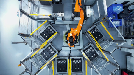 Top‑down view of an industrial automation cell with a robotic arm at the center. The robot is surrounded by multiple workstations arranged symmetrically, with metal frames, trays, and components organized around the handling area. Yellow safety markings are visible along the edges.