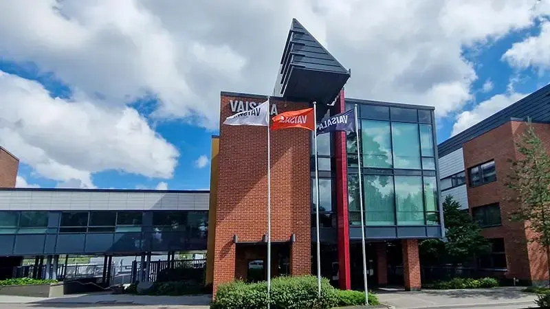 Exterior view of a modern brick and glass office building with vertical red accents and a triangular roof element. Flags are displayed in front, and the facade includes signage reading “VAIS.” The sky is partly cloudy above the surrounding structures.