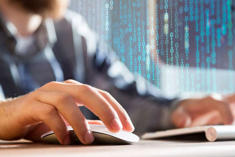 Close-up of a hand using a computer mouse on a desk. A keyboard and a person working at a computer are visible in the background. A digital overlay of binary code appears across the image.