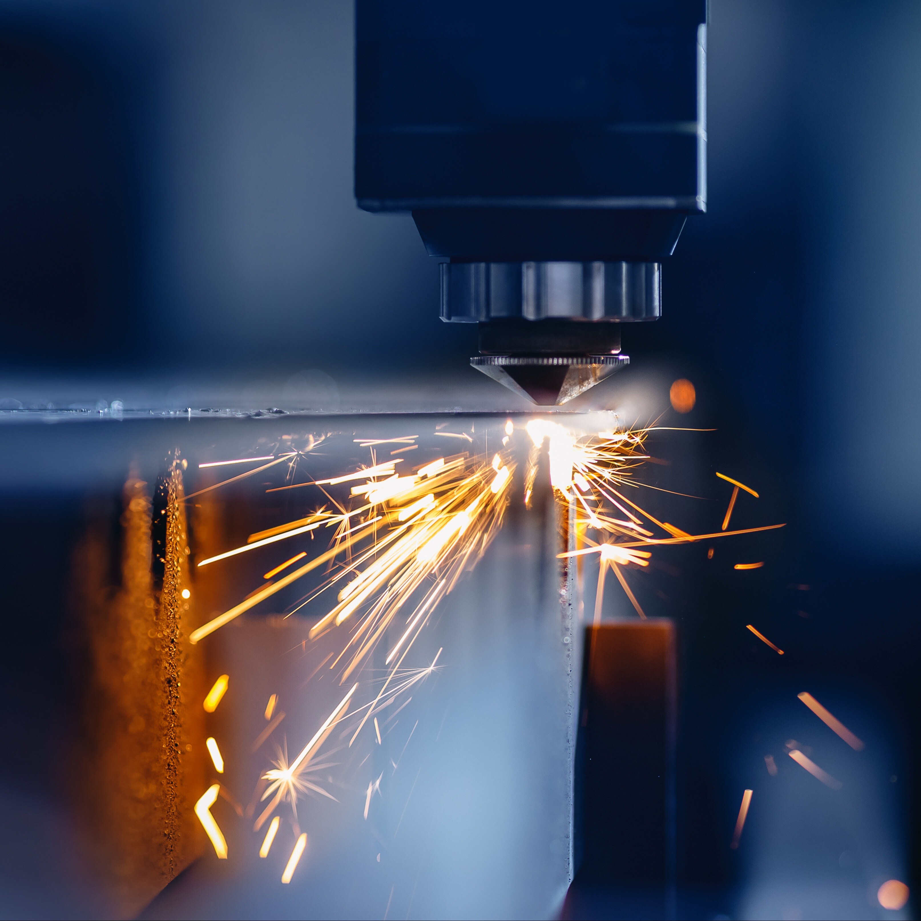 Close-up of a laser cutter working on a metal surface, with bright sparks emanating from the point of contact, against a blurred industrial background.