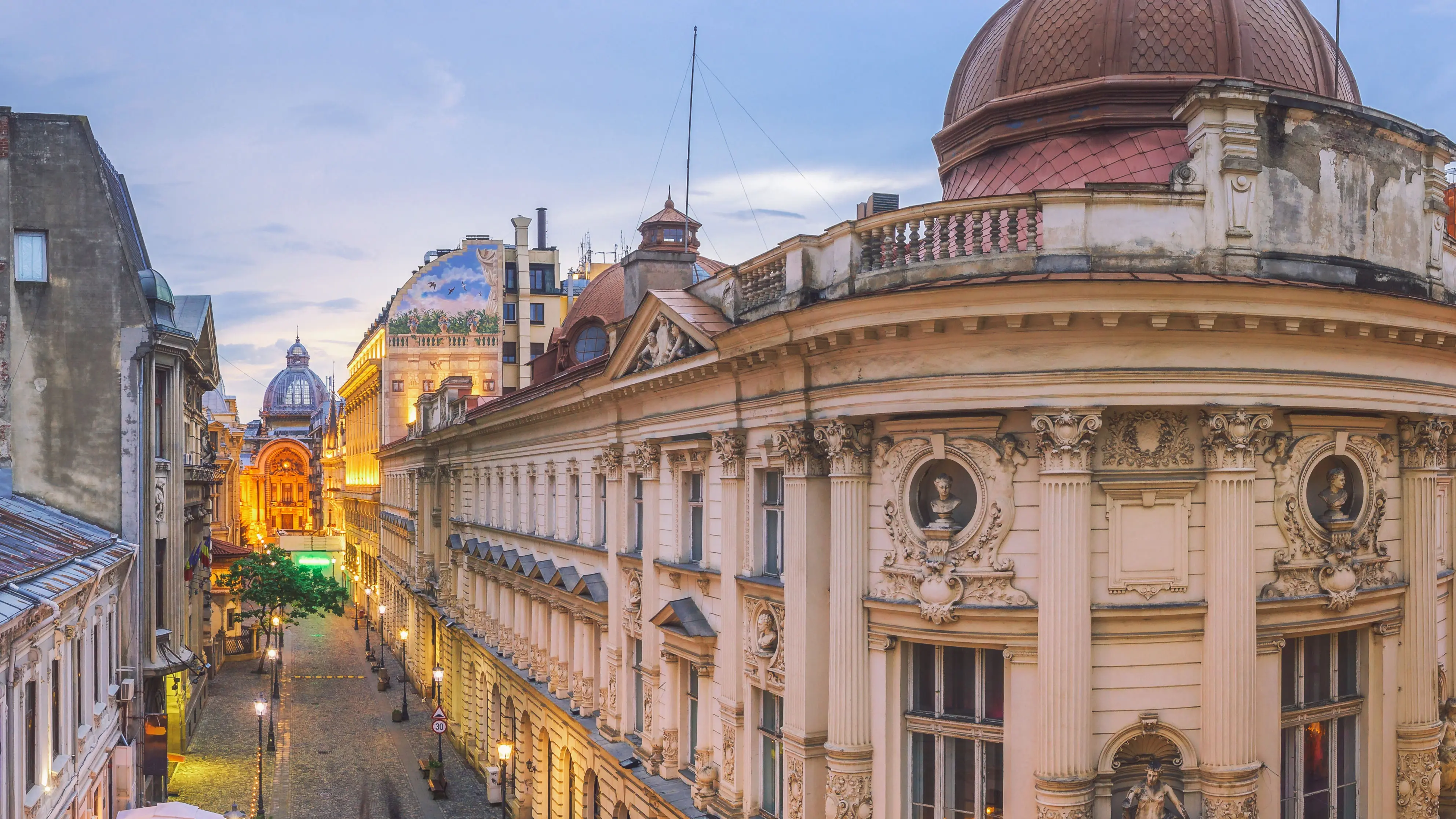 Narrow street in Bucharest lined with historic buildings featuring ornate façades, columns, and domed roofs. Warm lights illuminate the street, and a large domed landmark is visible in the background under a cloudy sky.