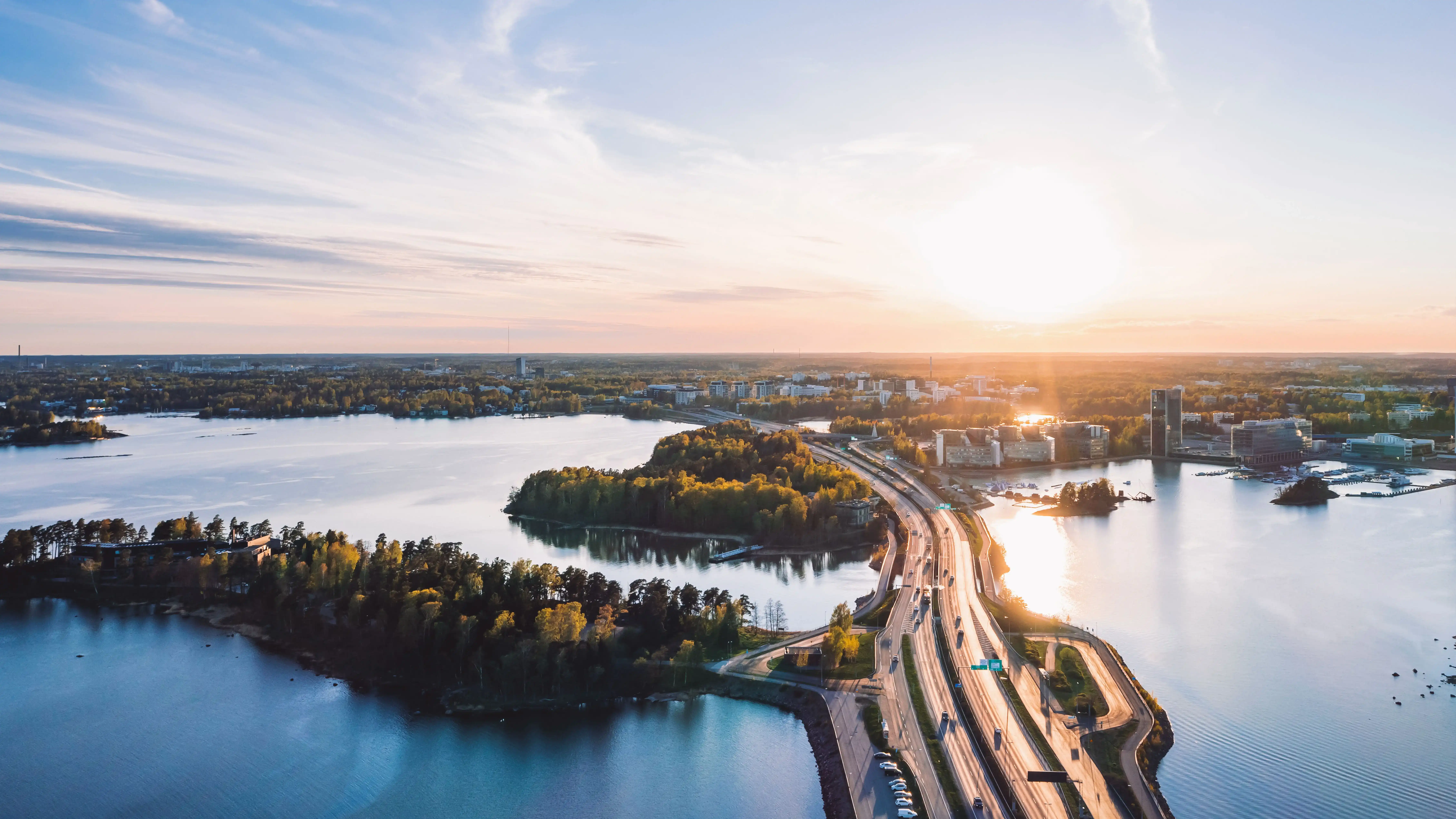 Aerial view of Espoo with a coastal landscape of water, small islands, and tree‑covered land. A roadway runs across the scene connecting the shoreline, with buildings visible near the water under a low sun and partly cloudy sky.