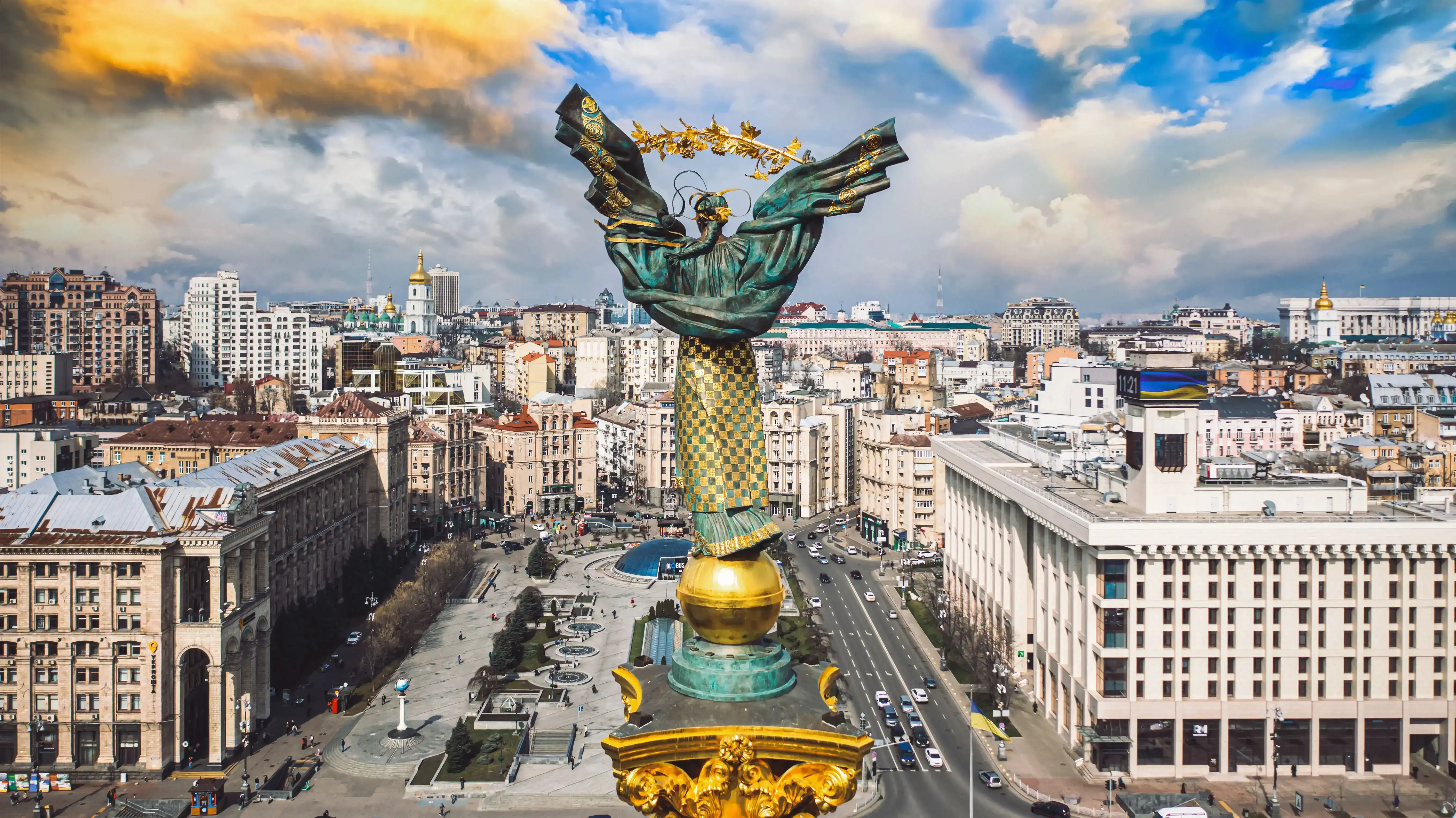 View of Kyiv featuring the Independence Monument at the center of Maidan Nezalezhnosti. The monument consists of a column topped by a statue holding a wreath, surrounded by city streets, buildings, and traffic under a partly cloudy sky.