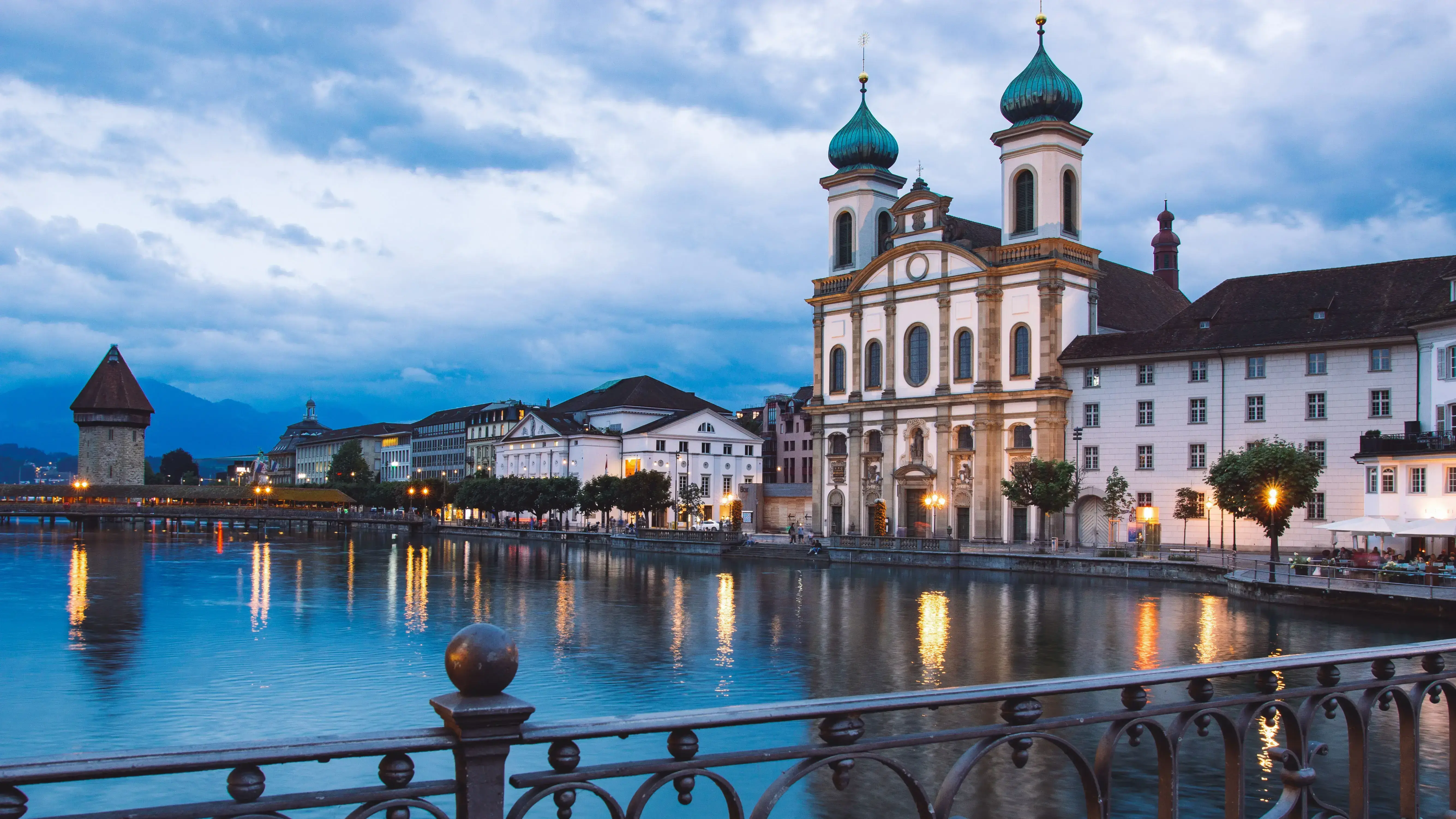 View of Lucerne along the river with historic buildings and the Jesuit Church featuring two towers. The Chapel Bridge and water reflections are visible, with mountains in the background under a cloudy evening sky.