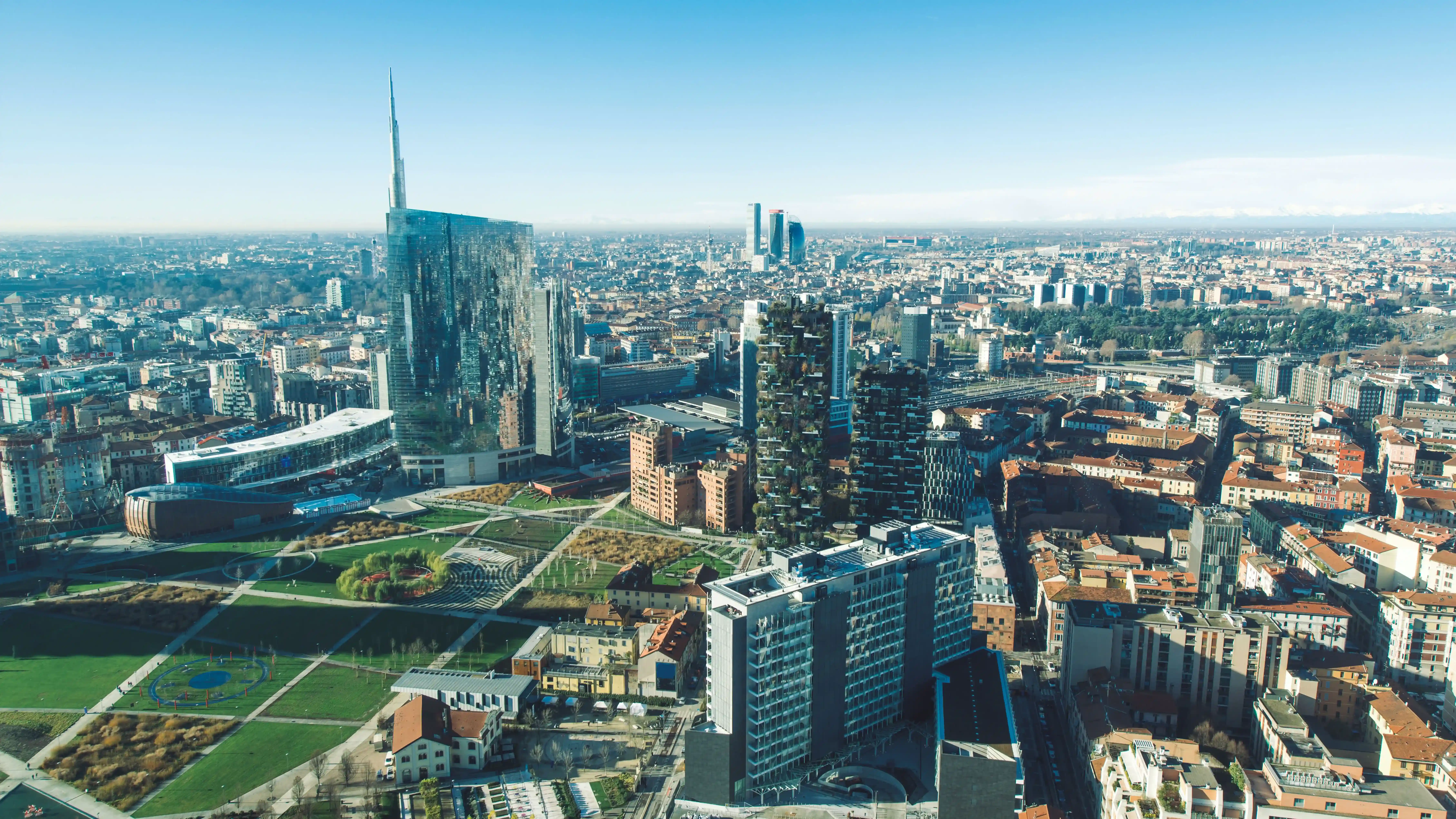 Aerial view of Milan showing a mix of historic and modern architecture, including high‑rise buildings and green terraces. Roads and city blocks extend across the landscape under clear daylight, with low hills visible on the horizon.